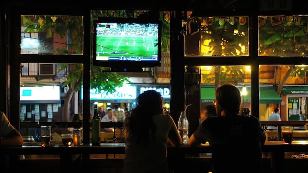 A photograph showing a crowded sports bar, with multiple large-screen TVs displaying different live sports games. The atmosphere is lively, with fans cheering and interacting, emphasizing the social aspect of watching sports in a public setting.