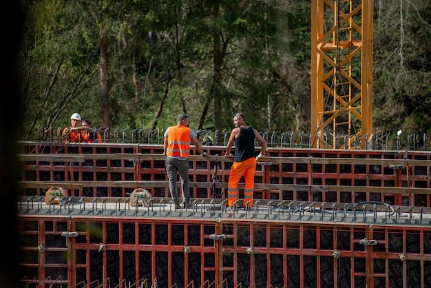 Construction workers in hard hats and safety vests on an active bridge construction site, with heavy machinery in the background, illustrating job creation and infrastructure development.